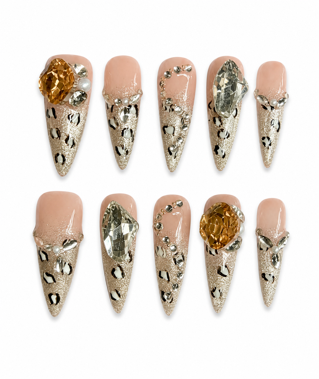 Luxury nude stiletto press-on nails with champagne glitter tips, leopard print accents, oversized crystal gems, pearl details, and glossy finish displayed on a white background.