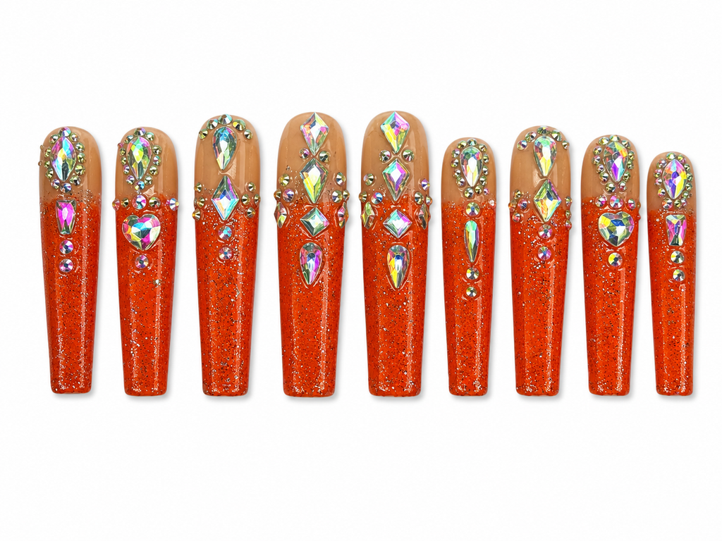 Long coffin press-on nails with nude and orange glitter ombré design, featuring holographic rhinestones and crystal embellishments on a white background.