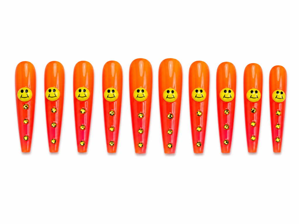 Extra-long coffin press-ons are bold orange-to-red ombre with a yellow smiley face charm on each nail and a trail of gold rhinestones cascading down the length. 