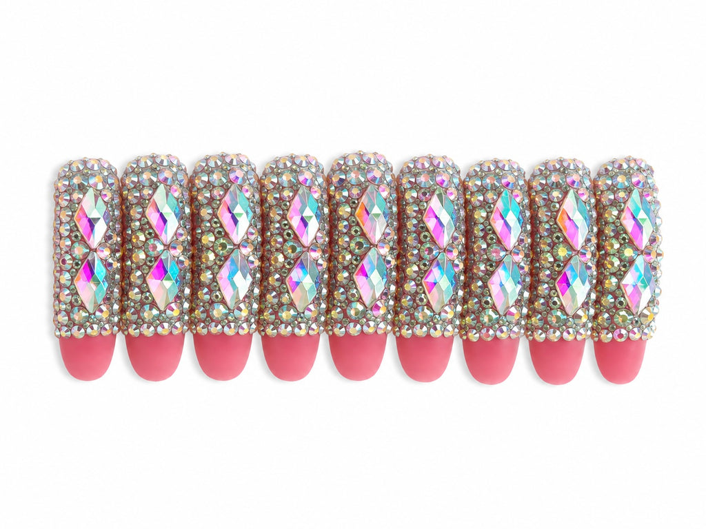Handmade luxury press-on nails featuring pink matte tips fully covered in iridescent rhinestones and oversized diamond-shaped crystal gems for a glamorous bling nail design on a white background.
