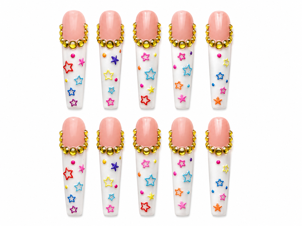 Handmade long coffin press-on nails featuring nude pink and white designs with gold rhinestone cuticle accents, colorful star art, and playful multicolor dot details on a clean white background.