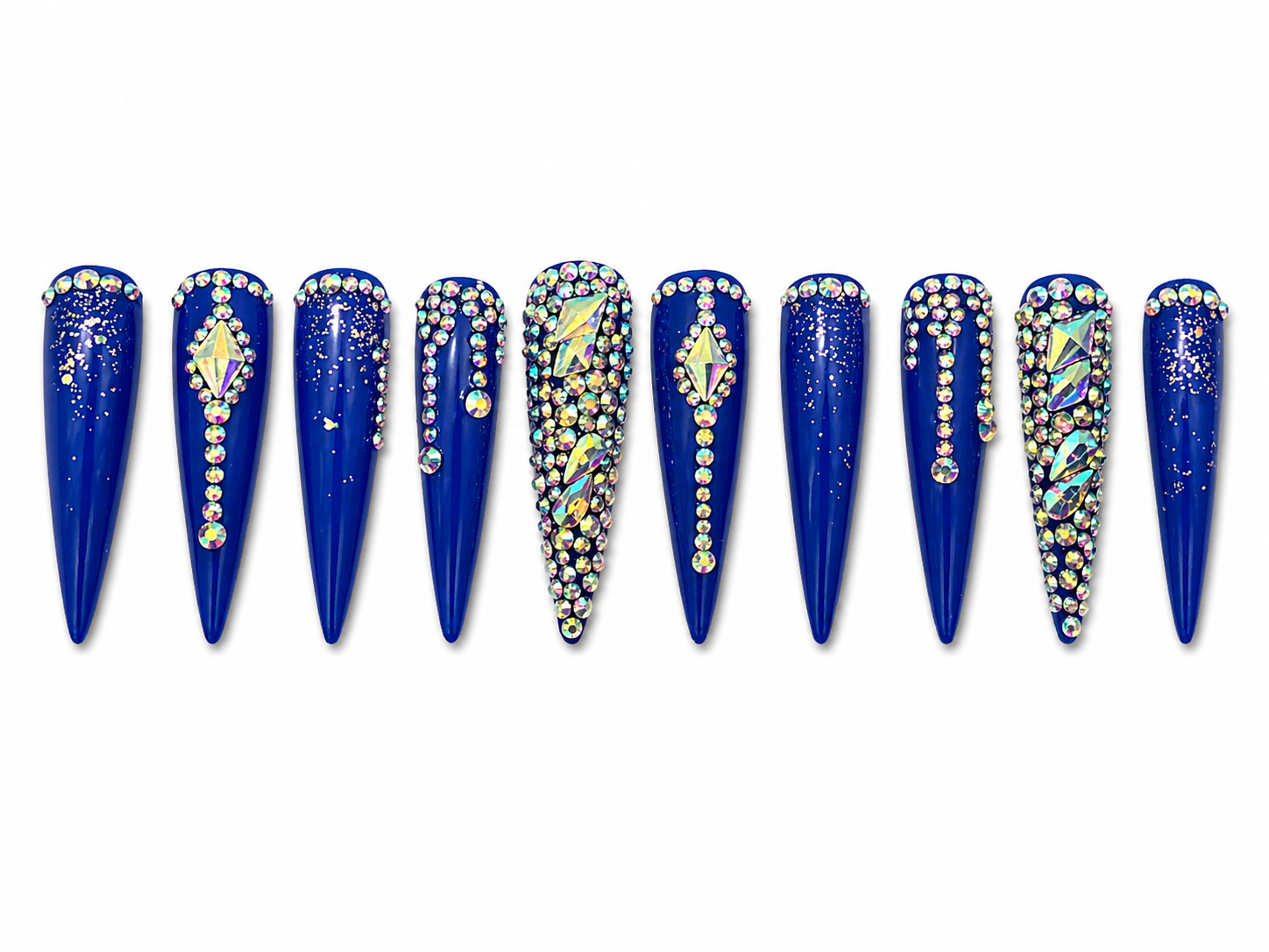 extra-long stiletto press-ons, royal blue — a rich, almost cobalt shade — covered in iridescent AB rhinestones arranged in arcs, trails, and dense clusters, with large diamond- cut gemstone centerpieces and scattered gold glitter on alternate nails. 